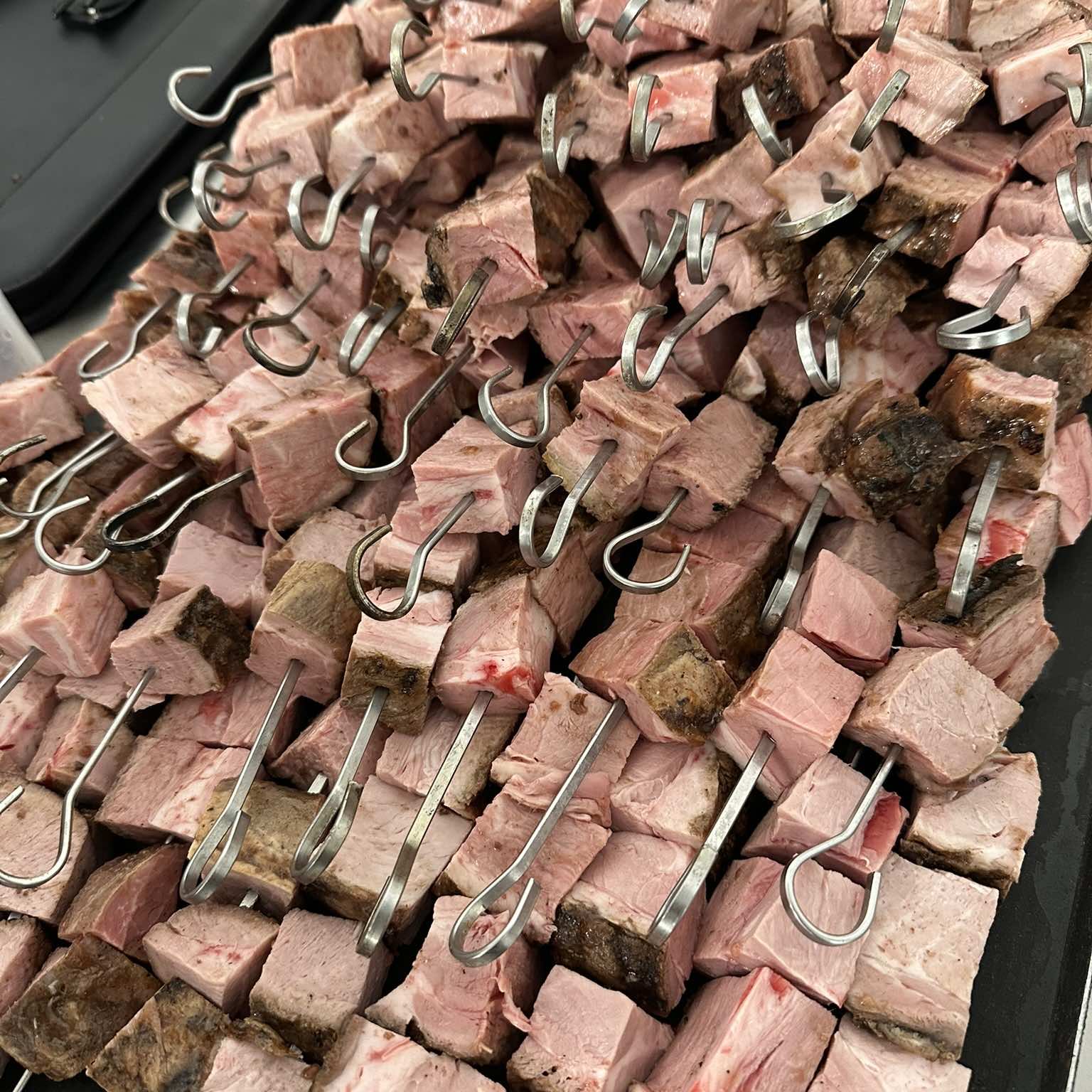 Cubes of braised beef brisket threaded onto metal skewers and piled on a tray, ready for the grill