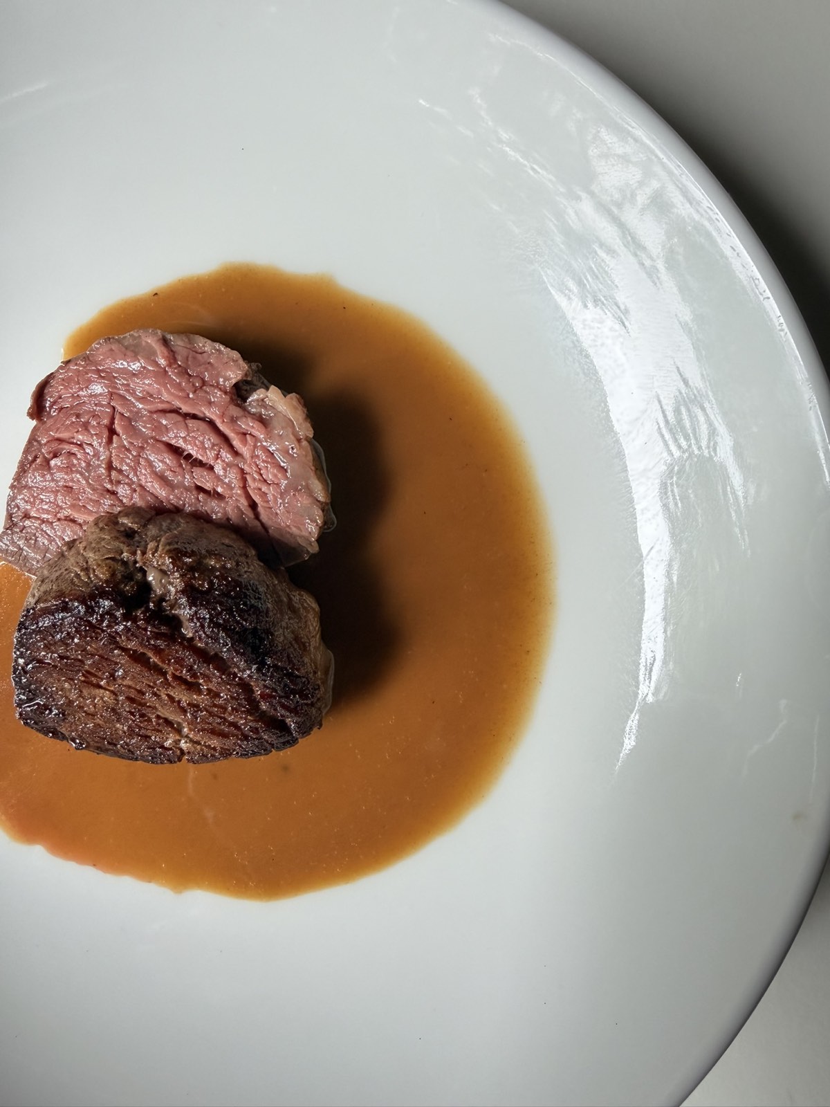 A seared eye fillet steak cut open to show the pink centre, sitting in a pool of glossy pan sauce on a white plate