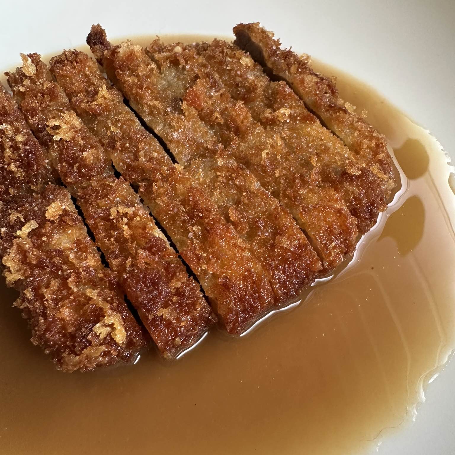 Panko-crumbed pork katsu, fried deep golden and sliced into strips, sitting in a pool of glossy jus gras in a white bowl